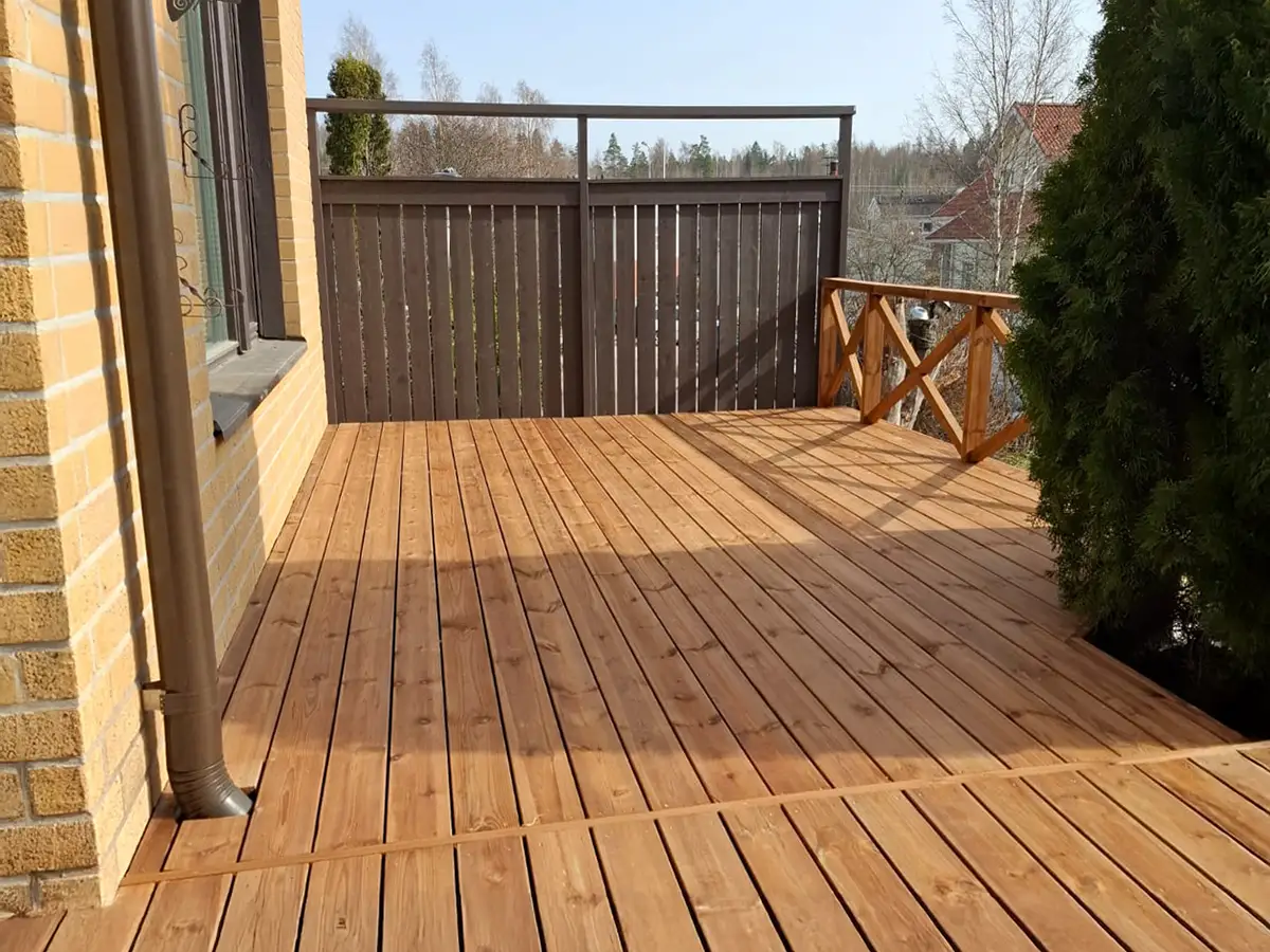 Puinen terassi pitkittäislaudoituksella ja kaiteella, sekä tummempi ruskea aitaelementti.