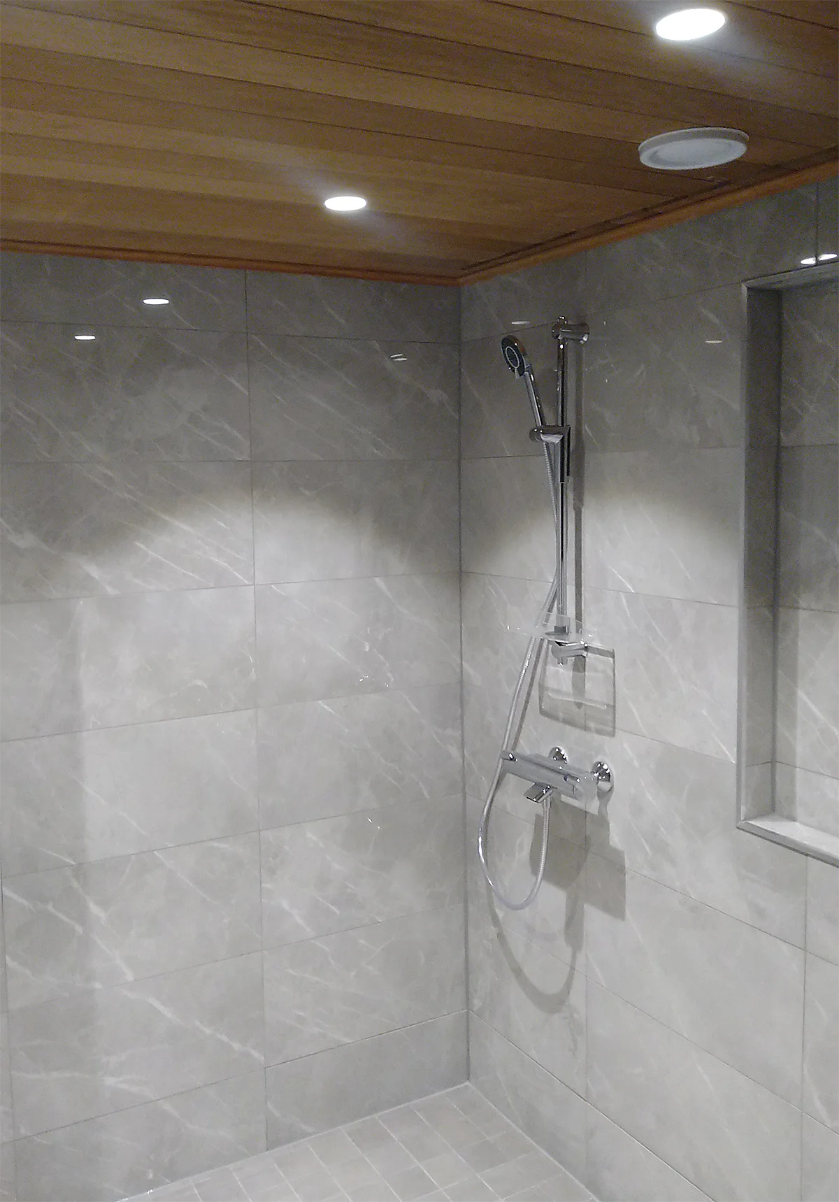 Kylpyhuoneessa vaalean harmaat laattapinnat, puupaneelinen katto ja suihkun vieressä seinäsyvennys pesuaineille.